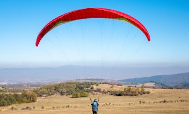 Vertical picture of a man flying with a red parachute surrounded by greenery under a blue sky.